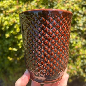 May include: A cylindrical ceramic tumbler featuring a deep brown, glossy glaze with a raised, repeating diamond-shaped texture across the exterior surface. The cup has a smooth, dark brown rim and a slightly tapered base, showcasing a reflective finish.