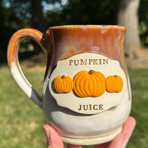Op de afbeelding: Een keramische mok met een handvat, een crèmekleurige basis en een bruine rand. De mok heeft een label met de tekst "PUMPKIN JUICE" en drie oranje pompoenontwerpen. De mok wordt door een persoon vastgehouden.