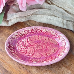 May include: A pink ceramic oval dish with a raised floral rose design. The dish is a vibrant pink color with a glossy finish. The background includes pink roses and a beige cloth on a wooden surface.