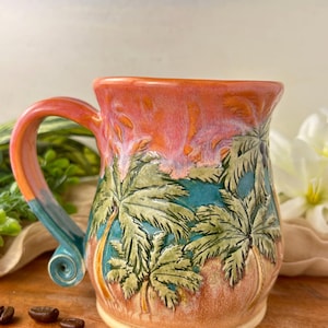 May include: A ceramic mug featuring a relief design of palm trees against a sunset sky. The mug has a gradient glaze transitioning from a warm coral pink at the rim to a deep teal blue at the base, with a curved handle ending in a small spiral.