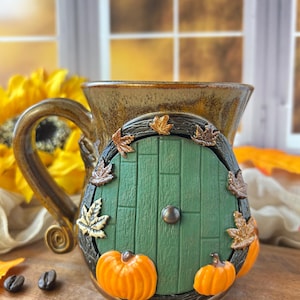 May include: A ceramic mug with a brown glazed finish featuring a sculpted green wooden-style door on the front. The door is surrounded by a dark textured frame adorned with small orange pumpkins and brown autumn leaves, with a central round knob.