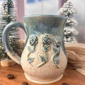 May include: A ceramic mug with a blue and cream glaze. The mug features three raised rose designs on the front. The handle and rim are blue. Coffee beans are scattered on the wooden surface in front of the mug.