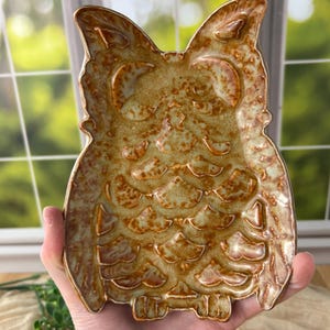 May include: A ceramic owl-shaped dish featuring a glossy, mottled brown and tan glaze with a crackled texture. The tray has raised, sculpted details representing feathers, pointed ears, and a rounded body, creating a distinct silhouette.