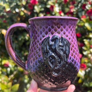 May include: A handcrafted ceramic mug with a dragon design. The mug features a textured, scale-like pattern in shades of purple and black. A black dragon is sculpted on the front. The handle and rim have a metallic sheen.