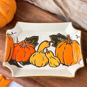 May include: Rectangular ceramic serving dish with a cream-colored base and a hand-painted design of orange and yellow pumpkins with green leaves. The dish has a scalloped edge and a brown border.