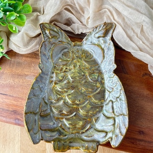 May include: A ceramic owl-shaped trinket dish featuring a textured surface with a metallic finish in shades of muted green, gold, and gray. The dish has detailed embossed feather patterns and defined owl facial features including large eyes and pointed ears.