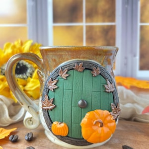 May include: A ceramic mug with a glossy brown glaze featuring a sculpted green circular door with vertical planks, a central knob, and brown autumn leaves. Two orange pumpkins are attached to the lower section of the door frame.