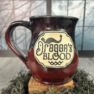 Pode incluir: Uma caneca de cerâmica com esmalte vermelho escuro e preto. A caneca apresenta um rótulo creme com as palavras "Dragon's Blood" em uma fonte decorativa. A caneca tem uma alça grande e está colocada em um pequeno bloco de madeira.