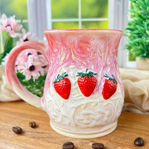 May include: A handmade ceramic mug with a pink and cream color scheme. The mug features three raised red strawberry designs with green leaves. The handle and rim are pink, and the body has a textured, floral pattern.