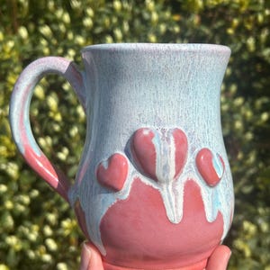 May include: A pink ceramic mug with a white glaze and three raised heart-shaped details. The mug has a light blue glaze that drips down the sides.