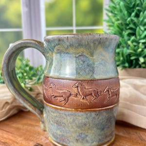 Peut inclure: Une tasse en céramique artisanale avec un motif de chevaux en relief. La tasse présente un glaçage bleu et marron, une grande anse et est remplie de grains de café. La tasse est posée sur une surface en bois.