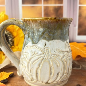 May include: A ceramic mug featuring a relief-carved pumpkin design on the lower half. The mug has a cream-colored base with a glossy, mottled olive-green and brown glaze dripping from the rim down to the top of the carved pumpkin pattern.