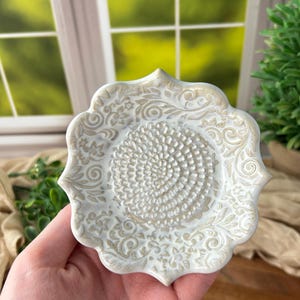 Puede incluir: Un plato rallador de cerámica circular con borde festoneado, que presenta un centro elevado y texturizado para rallar y un intrincado patrón floral y de enredaderas en relieve por toda la superficie en tonos blanco roto y beige claro.
