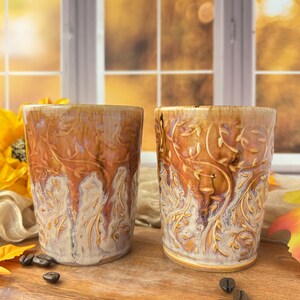 May include: Two ceramic tumblers featuring a raised, embossed vine and leaf pattern. The glaze transitions from a warm, glossy amber brown at the top to a creamy, textured off-white at the base, creating a distinct drip-like effect on the surface.