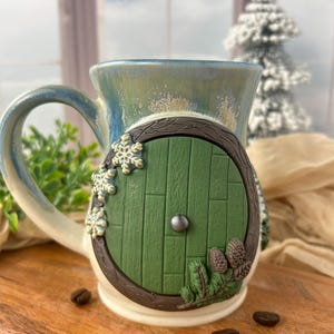 May include: A ceramic mug with a whimsical design featuring a green door with a silver knob, surrounded by brown trim, snowflakes, and pinecones. The mug has a blue and green glazed finish and a large handle. Coffee beans are scattered around the base.