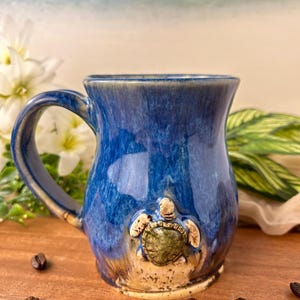 May include: A ceramic mug with a blue glaze and a sculpted sea turtle design. The mug has a curved handle and a wide mouth. Coffee beans are scattered around the base of the mug. The mug is approximately 4 inches tall.