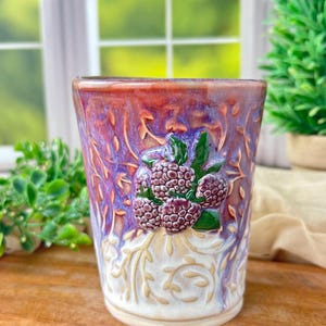 May include: A ceramic tumbler featuring a raised relief of purple raspberries and green leaves on the front. The surface has a blended glaze of deep red, purple, and white, with an embossed vine pattern covering the lower portion of the vessel.