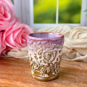 May include: A ceramic tumbler featuring a relief-carved rose pattern. The vessel transitions from a deep brown base with carved floral details to a smooth, glossy purple and pink rim. The surface texture highlights the intricate rose motifs.