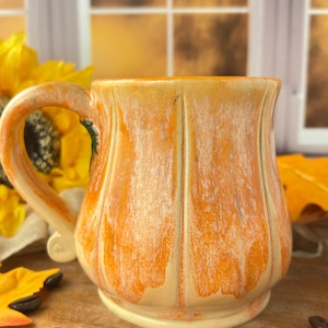 May include: A ceramic mug shaped like a pumpkin with a glossy orange and cream glaze. The mug features vertical ridges mimicking the texture of a gourd, a curved handle on the left side, and a wide base. The finish shows a blended color gradient.