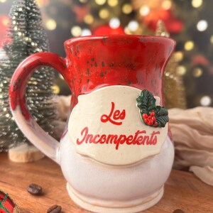 May include: A handmade ceramic mug with a red speckled top and handle, transitioning to white. The mug features a cream-colored label with the words "Les Incompetents" in red, and a holly accent. Coffee beans are scattered around the mug.