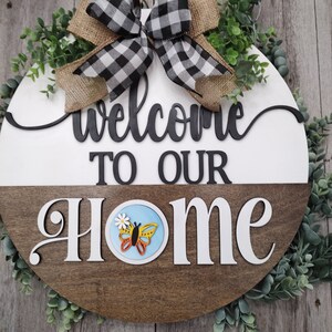 Peut inclure: Une pancarte ronde en bois de bienvenue avec un fond blanc et brun. La pancarte dit "Bienvenue chez nous" en lettres noires et blanches. Un nœud à carreaux noir et blanc avec un ruban de toile de jute est en haut de la pancarte. Des brindilles vertes feuillues entourent la pancarte.