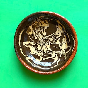 May include: A handmade ceramic bowl with a brown exterior and a cream and brown swirled interior design. The rim is a reddish-brown color. The bowl is set against a bright green background.