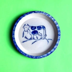 May include: A round ceramic plate with a blue and white cow design. The plate has a blue patterned border and a white center with a blue cow illustration. The cow has blue spots and is standing on a blue line.