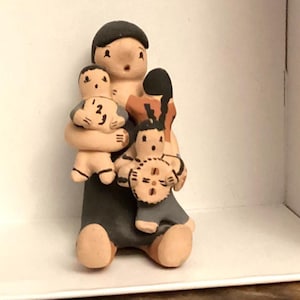 May include: A ceramic figurine depicting a person holding two children. The figures are painted in muted tones of tan, black, and gray. One child holds a clock, and the other holds a circular object. The figurine is displayed in a white box.