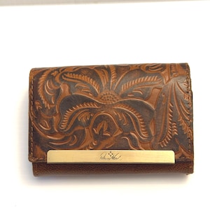 May include: Brown leather wallet with embossed floral design. Features a brass-colored metal bar with the brand name "Patricia Nash" engraved. The wallet is closed and has a rectangular shape.