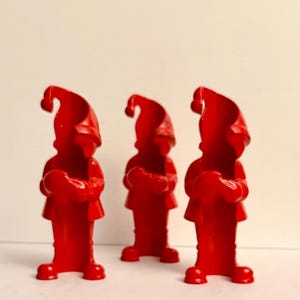 May include: Three red gnome figurines. Each gnome is wearing a tall hat and holding a heart-shaped object. The figurines are standing upright and have a glossy finish. The background is a plain, off-white color.