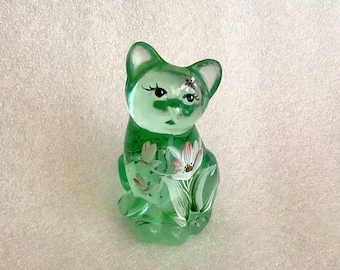 May include: A translucent green glass cat figurine. The cat has painted details, including eyelashes and floral accents. The figurine is sitting upright with its paws together, and the overall design is delicate and detailed.