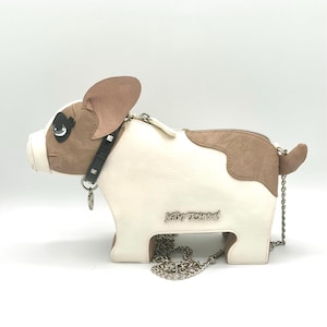 May include: A crossbody bag shaped like a French bulldog in cream and tan. It features a black studded collar with a silver charm, a zipper closure, and a silver chain strap. The text Betsey Johnson is embossed in silver on the side of the bag.