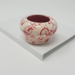 May include: A small, white ceramic planter with a red and white design. The planter has a rounded shape and a wide opening. It is decorated with a pattern of red flowers and leaves.
