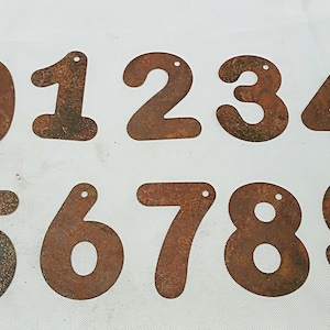 Può includere: Dieci numeri in metallo arrugginito, da 0 a 9, con fori per l'appeso. Ogni numero è tagliato individualmente e ha una forma unica, leggermente irregolare.