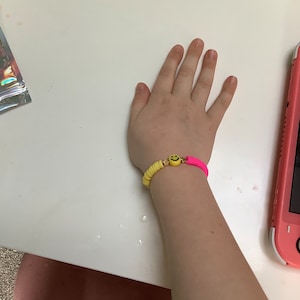 Puede incluir: Un brazalete de cuentas rosa y amarillo con un dije de carita sonriente. El brazalete está en la muñeca de una persona.
