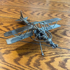 Puede incluir: Modelo de avión biplano de metal hecho a mano con un motor y hélice detallados. Las alas y el cuerpo son de metal, y el modelo se encuentra sobre una superficie de madera. El biplano tiene una estética vintage e industrial.