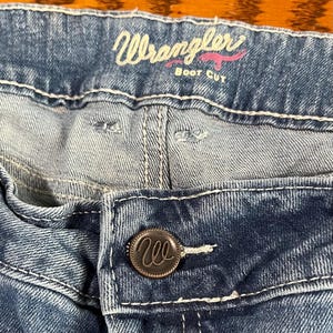 May include: Close-up of blue denim jeans with white stitching. The jeans feature a button closure with a circular metal button. The word "Wrangler" is embroidered in pink script above the front pocket, with "Boot Cut" underneath.