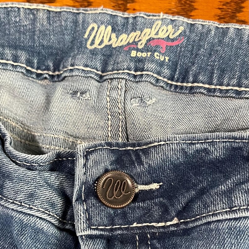 May include: Close-up of blue denim jeans with white stitching. The jeans feature a button closure with a circular metal button. The word "Wrangler" is embroidered in pink script above the front pocket, with "Boot Cut" underneath.