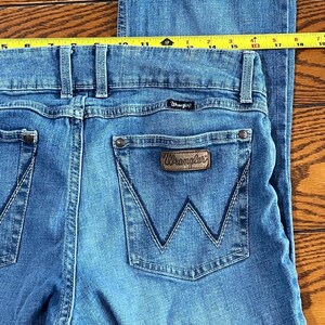 May include: Close-up of blue denim Wrangler jeans. The jeans have a classic five-pocket design with a brown leather patch on the back pocket. A yellow measuring tape is placed across the top of the jeans. The jeans are a light wash.