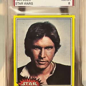 Könnte beinhalten: Eine bewertete Star Wars Sammelkarte mit Han Solo aus der Topps-Serie von 1977. Die Karte ist in nahezu neuwertigem Zustand und trägt die Nummer #139. Die Karte zeigt ein Nahaufnahmebild von Han Solo mit dem Text "Han Solo... Held oder Söldner?" und dem Star Wars-Logo.