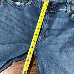 May include: Close-up of blue denim jeans with a measuring tape showing a length of approximately 11 inches. The jeans have a button closure and visible stitching details. The fabric has a faded appearance.