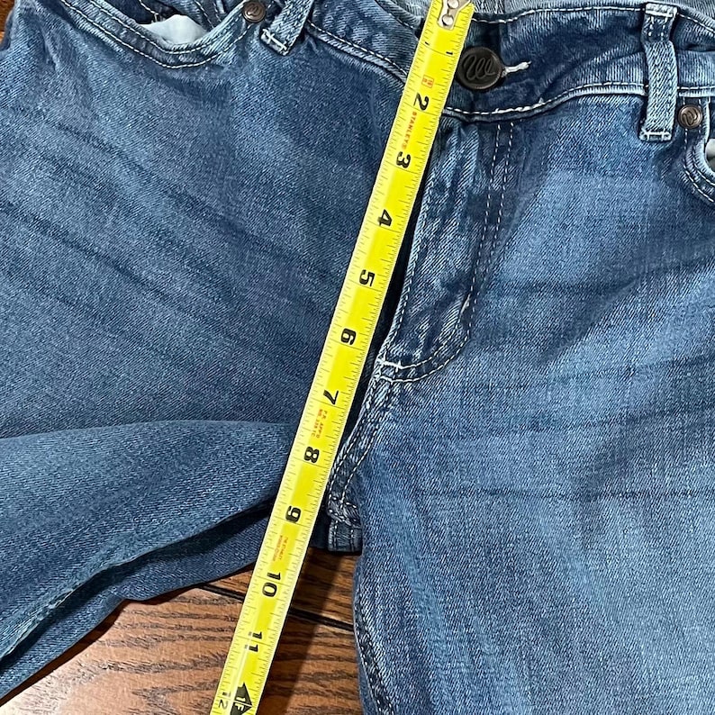 May include: Close-up of blue denim jeans with a measuring tape showing a length of approximately 11 inches. The jeans have a button closure and visible stitching details. The fabric has a faded appearance.