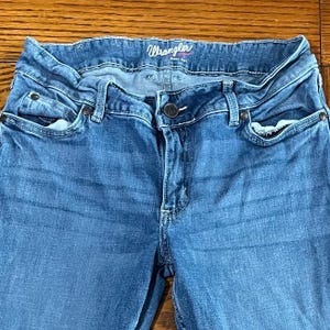 May include: Close-up of blue denim Wrangler jeans. The jeans have a classic five-pocket design, a button and zipper closure, and belt loops. The Wrangler logo is visible on the waistband.