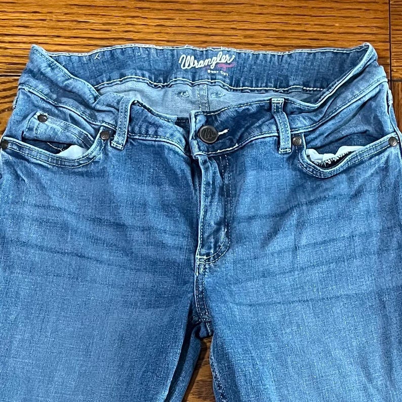 May include: Close-up of blue denim Wrangler jeans. The jeans have a classic five-pocket design, a button and zipper closure, and belt loops. The Wrangler logo is visible on the waistband.