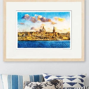 May include: A framed watercolour print depicting a coastal city with a domed church and a tall spire, set against a blue sky with fluffy clouds. The print is in a light wood frame, above a white cushioned bench with decorative cushions.