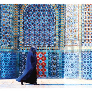 Puede incluir: Una mujer con un pañuelo azul camina frente a una pared cubierta de intrincados azulejos de mosaico azul, rojo y amarillo.
