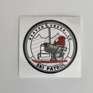 May include: Black and white sticker with a cartoon beaver wearing a red ski patrol hat and holding a bone. The beaver is riding a ski lift and the sticker says "Beaver Creek Co. Ski Patrol".