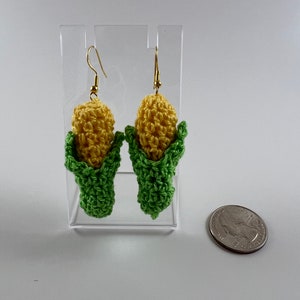 May include: A pair of crocheted corn on the cob earrings with green husks and yellow kernels. The earrings are hanging from gold hooks and are displayed on a clear acrylic stand.