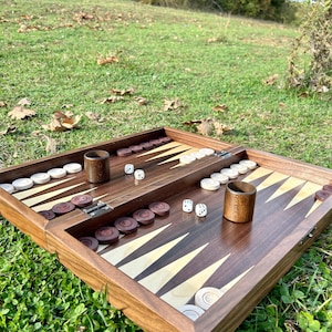 May include: A wooden backgammon set with a dark brown playing surface, featuring alternating light and dark triangles. The set includes brown and white game pieces, two wooden dice cups, and dice. The game is set up on a green grassy surface.