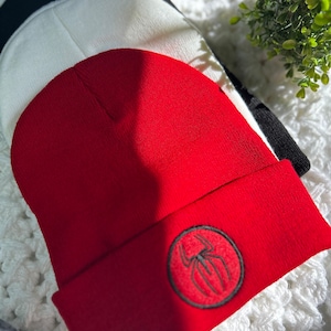 May include: A red beanie with a black embroidered spider logo. The beanie is folded and sitting on a white fuzzy surface.